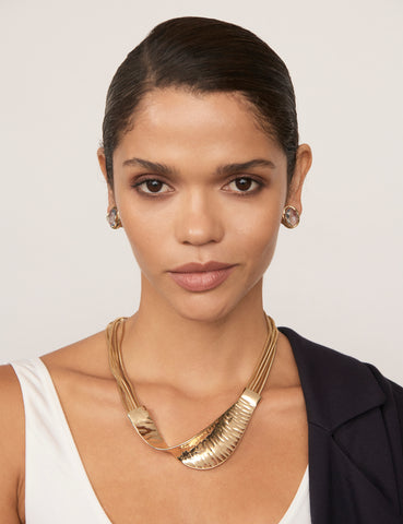 A woman with tan skin and dark hair wearing the Tempest Necklace. It features a contoured collar in gold-coloured plating. She wears a white singlet and a black blazer on one shoulder.
