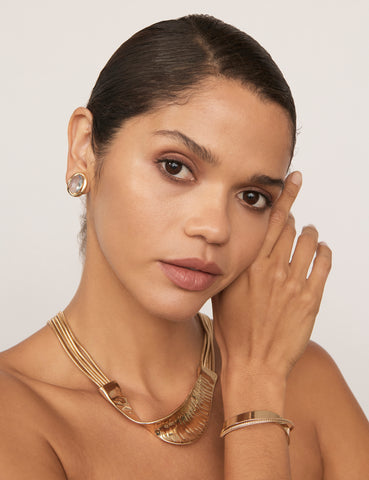 A woman's hand near her face wearing the Tempest Bangle. It is a solid gold-coloured bangle with two crossed bands. She also wears a matching gold necklace and earrings.