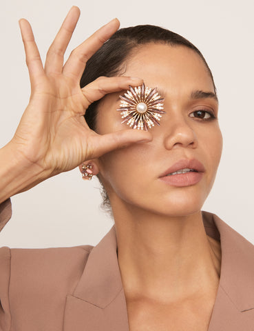 A woman holds the Dandelion Pearl Brooch in front of her eye. The brooch features a starburst design with gold-coloured plating, a faux pearl accent, faceted red stones and sparkling details. The woman wears a tan blazer and has dark hair.