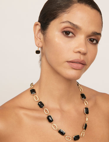 A woman wearing gold black jewellery, including a gold and black stone necklace and matching black stone earrings. The woman is tanned with dark hair and eyes.