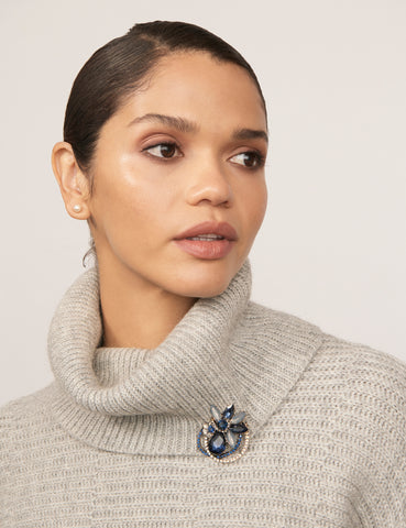 A woman wearing a grey turtleneck sweater with the Coil Petal Brooch. The brooches features mixed faceted blue and white stones with fine sparkling detailing. The woman has tan skin and dark hair.
