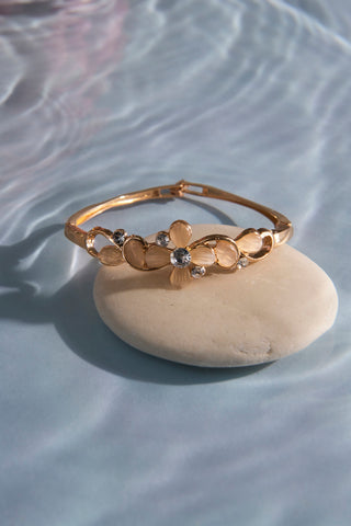 The Illume Blossom Bangle rests on a white stone under clear water. It is a gold-coloured bangle with natural-toned stones in a flower design and fine sparkling details. Water ripples gently against a blue background.