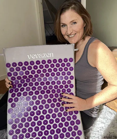 A woman smiles at the room as she holds a yoga mat marked 'vevazen,' which features a purple disc pattern. She looks happy and satisfied as she displays the mat, and the home environment around her suggests a moment of relaxation or preparation for a wellness exercise.