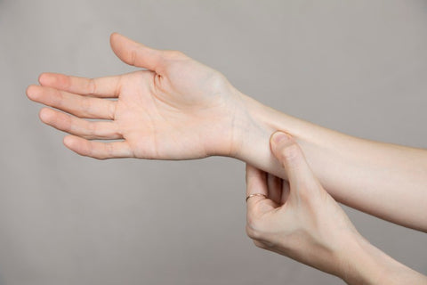 A woman is performing acupressure on her wrist, applying pressure with her thumb to point PC6, which is three fingers below the point where the wrist meets the palm. The background is neutral and highlights the therapeutic action.