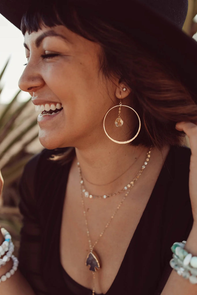 Woman smiling wearing gold hoop earrings with a citrine crystal dangling inside. 