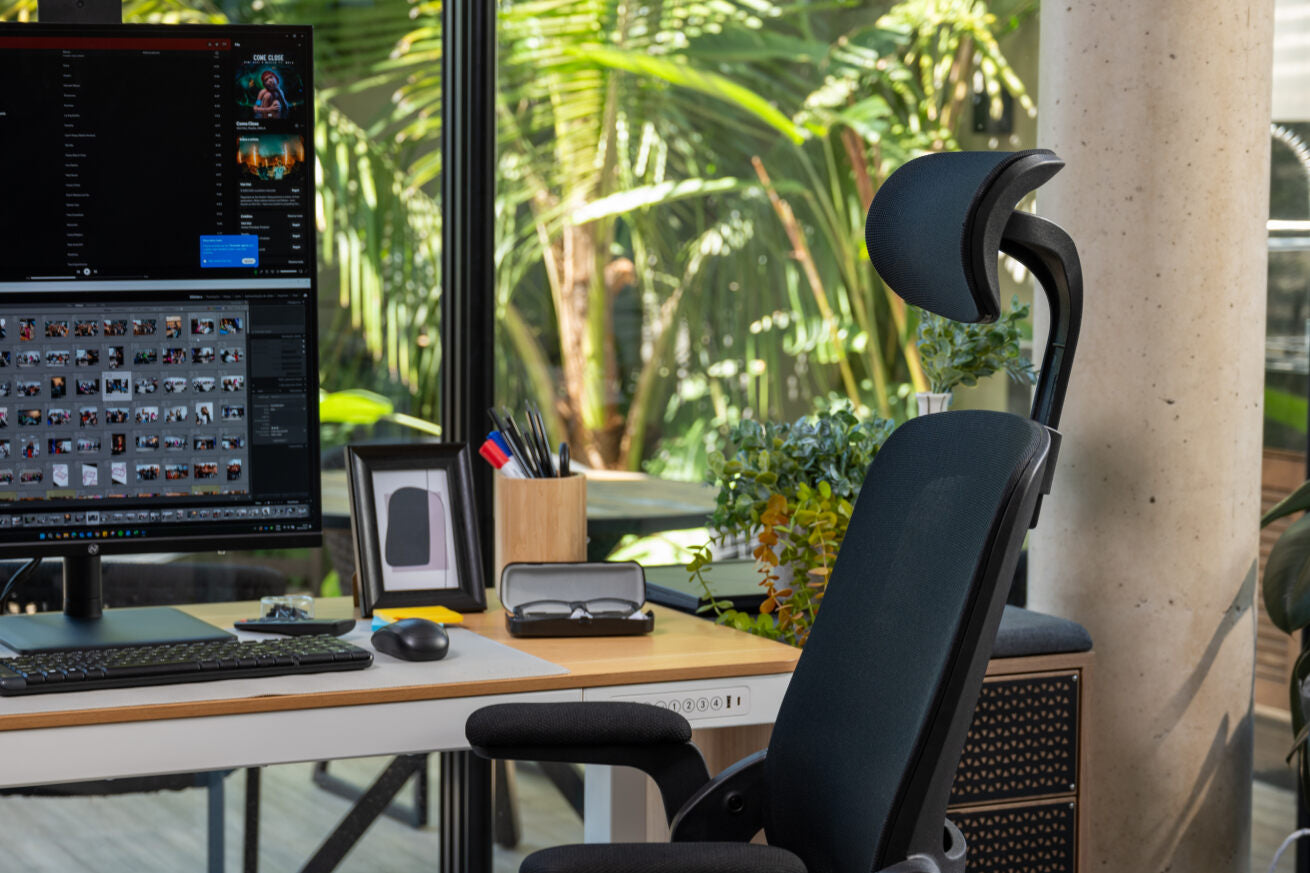 Escritório com plantas, monitor vertical, cadeira ergonômica e mesa elétrica com ajuste de altura.