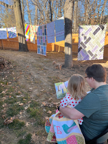 Birdie sitting in Nana's lap reading The Quilt Garden with quilts hung up around them