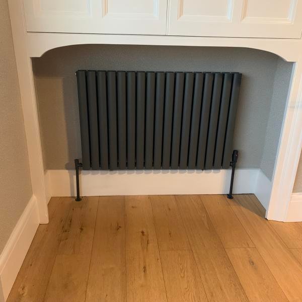 An Omeara black horizontal radiator from UK radiators sits within framing white woodwork beneath white cabinets.