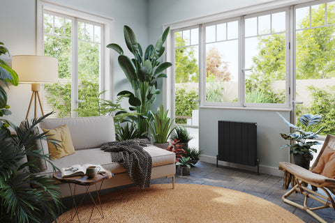 A black aluminium panel radiator against a pale blue wall in a bright living room full of tropical plants and cosy furnishings.