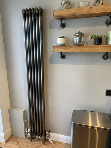 A slim vertical column radiator in a natural metal finish is pictured in a customer’s kitchen against a soft pearl grey wall.