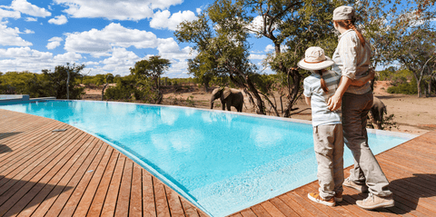 A mother a daughter stand by a swimming pool at a safari lodge, watching and elephant graze