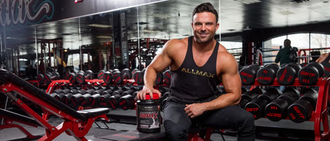 A muscular Allmax athlete holds a container of Isoflex protein in a gym.