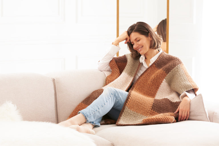 Women Wearing Caron Crochet Colorblock Ruana, Siting on Sofa with a smile on her face