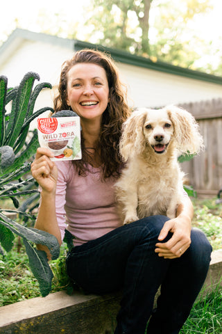 A woman, Zora, sitting with her dog, smiling as she holds up a package of Chili Beef Meat and Veggie Bar