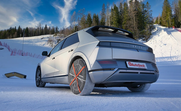 AutoSock snow socks mounted on rear wheels of a gray Hyundai Ioniq 5 car, standing on snow in skiing area