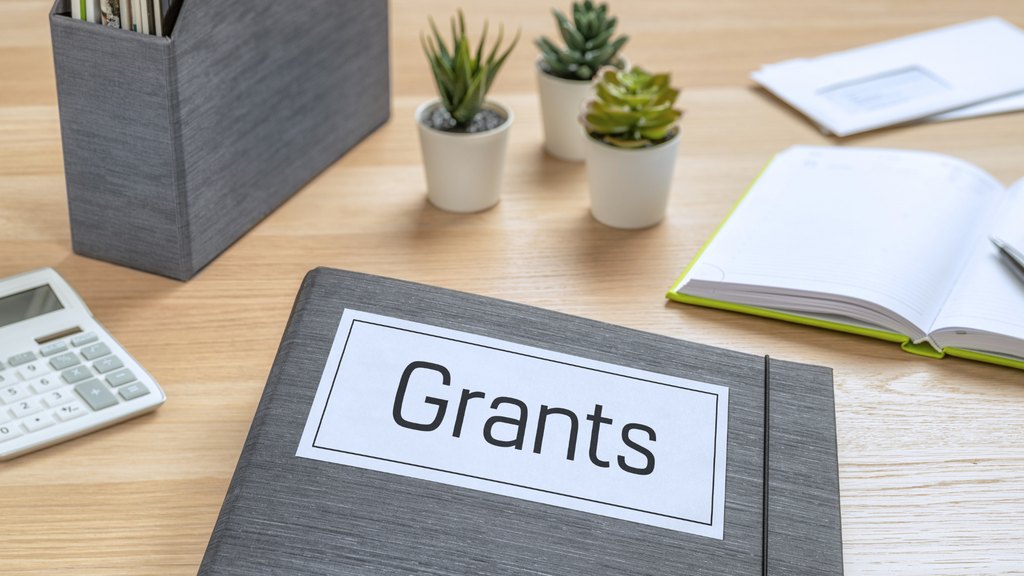 Folder printed with text "Grants" sitting on top of desk with some accessories
