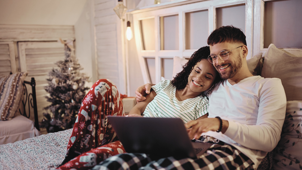 couple in christmas pajamas sitting on bed video conferencing with loved ones