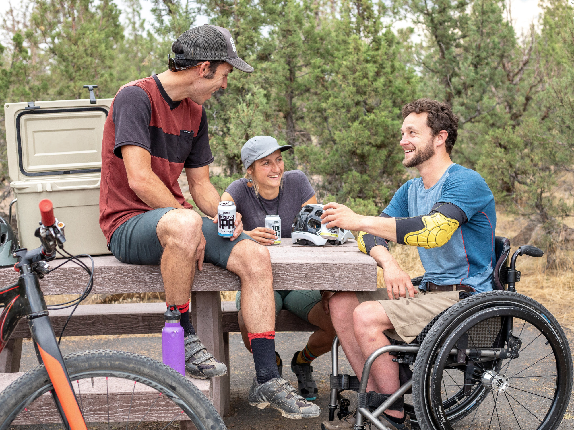 Three people drinking beer at a picnic table. They have been cycling