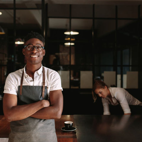 A smiling barista with his arms folded, wearing an apron with a woman working behind him.