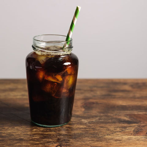 A glass of black cold brew coffee with a green and white paper straw in it, on a dark stained wood table.