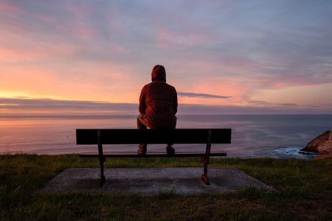   Lonely man in a hoodie sits alone on the rocky coast and enjoying sunset over rocky cliff to ocean horizon.