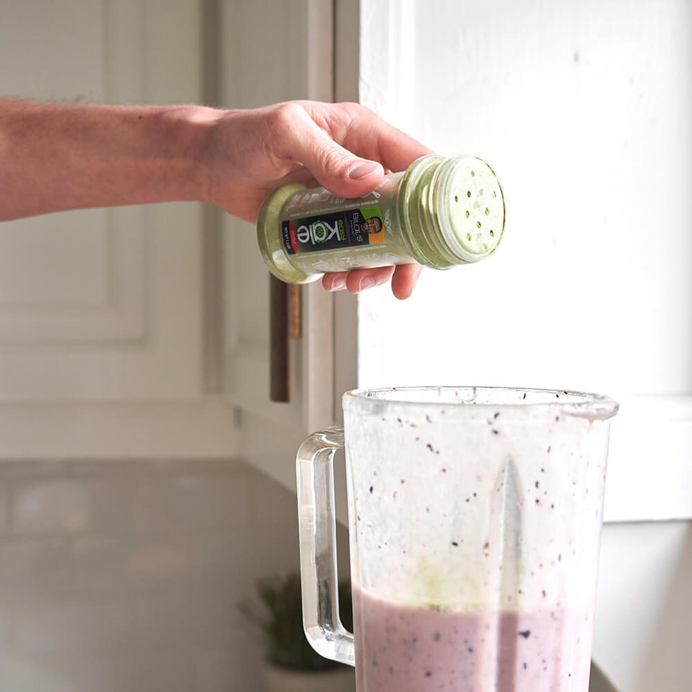 A man's hand sprinkles some of Bilal's EasyKale kale shaker into a nutritious smoothie within a blender cup