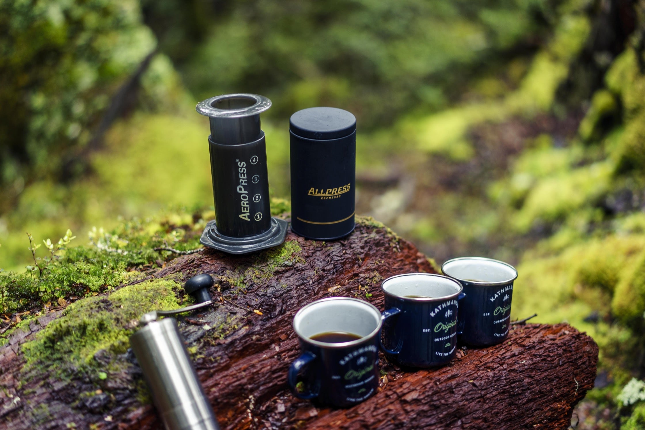 An Aeropress and other coffee accessories on a mossy tree log, with three mugs of black coffee.