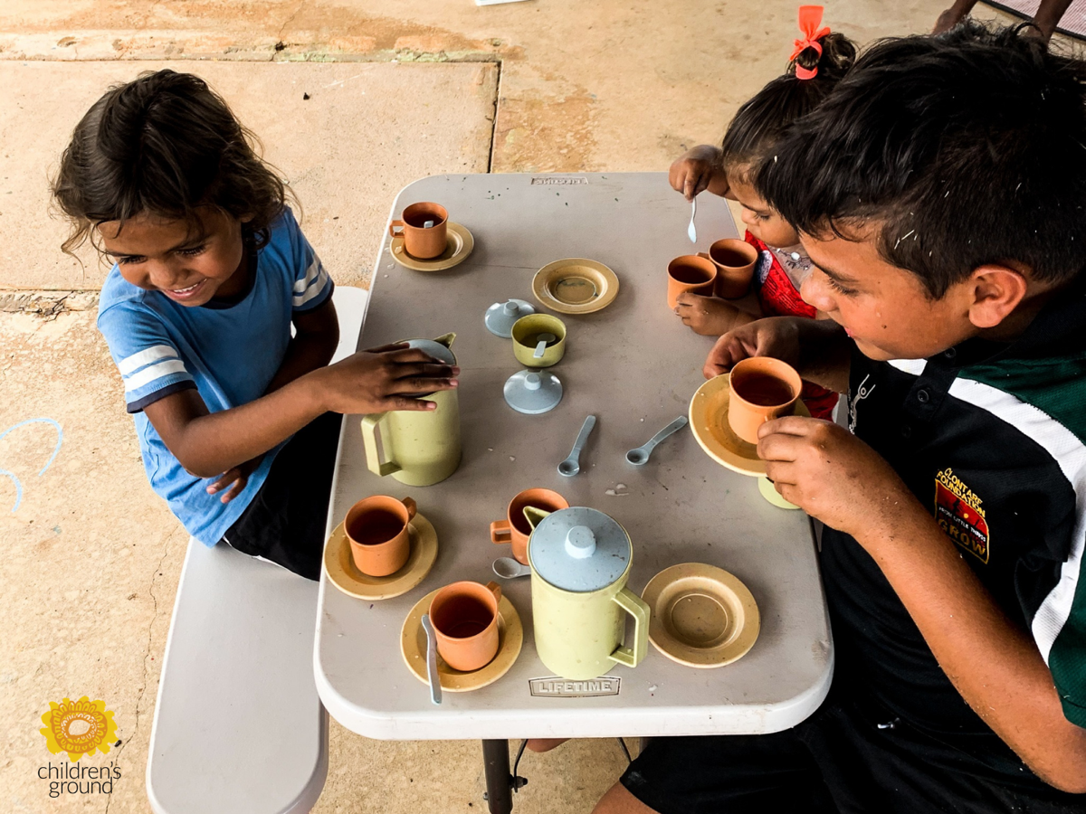 Three First Nations children enjoy a tea (coffee?) party. Photo courtesy of Children's Ground.