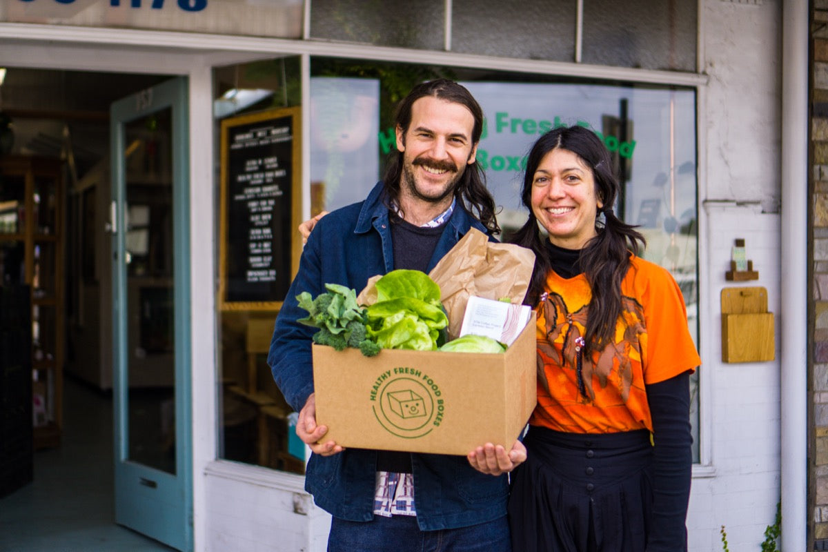 A portrait of Andrew and Minna outside the Brunswick West Workshop. Andrew holds a Health Fresh Food Box containing leafy greens and Sow coffee.
