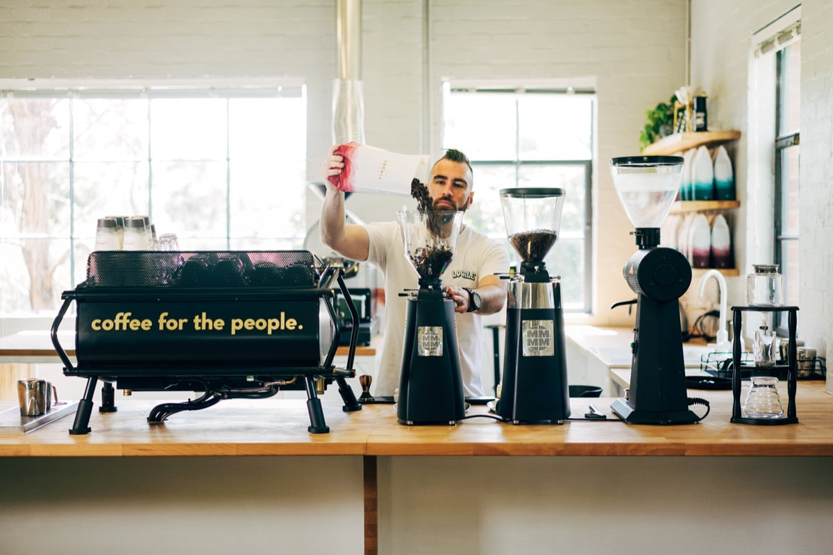 A member of the Locale Coffee Roaster team pours coffee beans into the hopper of a grinder. The coffee machine on the bench bears the slogan 'Coffee for the people.'