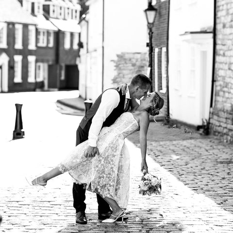 Black and White image of young couple in wedding attire on cobble street. Bride is holding a bouquet. Groom is holding brides leg up as she leans back and they kiss