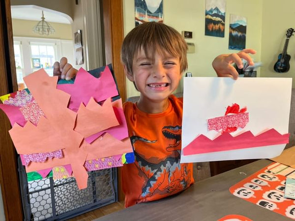 A boy holds up two of his artworks -- one a mixed media construction paper collage, and one a red drawing of mountains and an aircraft.