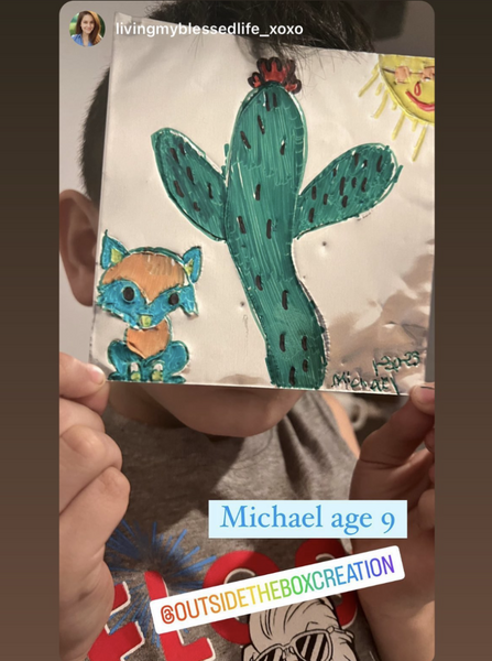 A boy holds up a small drawing of a blue and orange fox under a large Saguaro cactus.