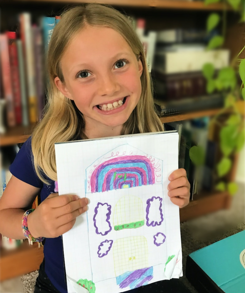 A young girl with blonde hair, a dark blue shirt, and a friendship bracelet on her arm proudly holds up her piece of art - a drawing of a house with a purple, pink, and blue roof.
