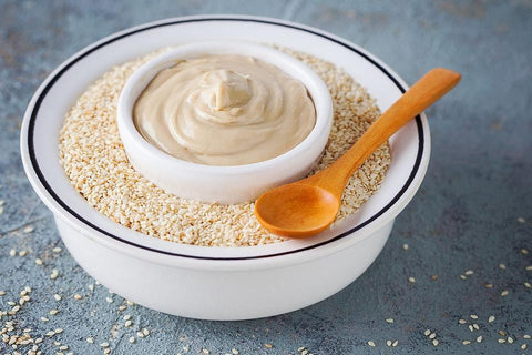 A rustic tableau unfolds: a bowl brimming with sesame seeds, crowned with a dollop of creamy tahini paste, complemented by a wooden spoon poised for a delectable journey into nutty richness.