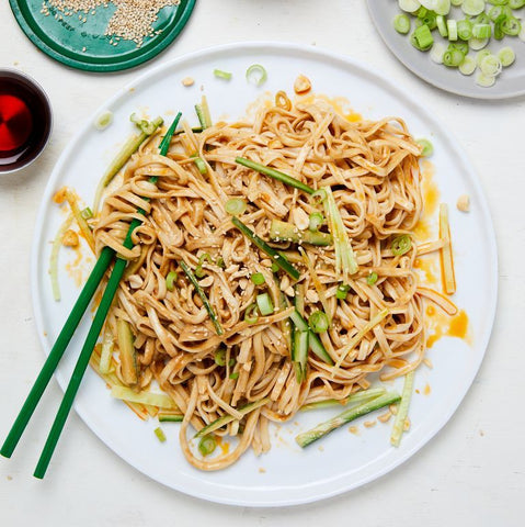 Asian noodles with tahini sauce, cucumber slices, spring onions, sesame side dish, and sprinkled sesame seeds.