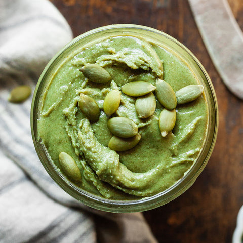 Indulge in the vibrant hues and flavors of pumpkin seed butter showcased in a glass jar, adorned with a garnish of green pumpkin seeds. A tantalizing blend of nuttiness and freshness awaits your culinary exploration.