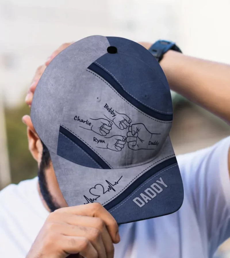Man holding a customized cap with handprints labeled as family members and the word 'DADDY' embroidered on it.