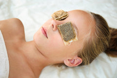 woman lying on a white pillow with a white towel over her chest and tea bags on her eyes