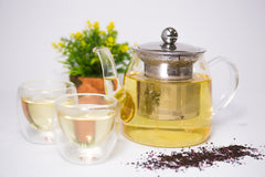 Image of glass teapot with silver infuser containing a light yellow colored tea with two clear cups of the same tea to the side, a plant behind and some looseleaf tea scattered in front.