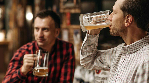 2 חברים יושבים לבירה בבר כשאחד מהם שותה את הבירה שלו מכוס זכוכית