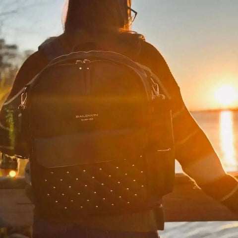 women wearing black backpack diaper bag with rose gold studs watching sunsetting over water