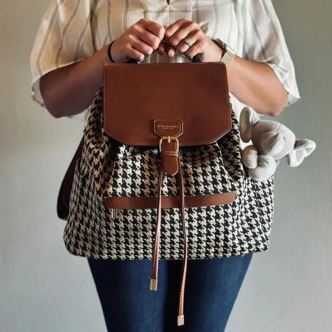 Woman holding a houndstooth diaper bag drawstring backpack with leather details and a baby's elephant toy hanging out the side pocket