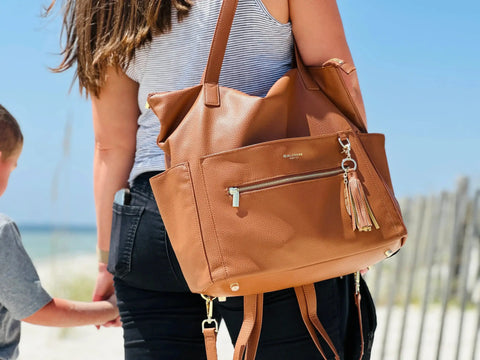Woman holding hands with children on the beach wearing brown leather diaper bag with leather and gold feather tassel by the shoulder straps