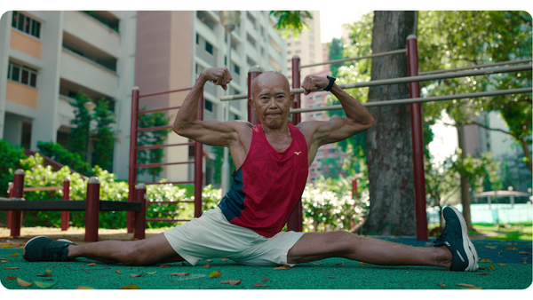An elderly man of Asian nationality, doing the splits and flexing his arms in a park with an apartment block and tree behind him.