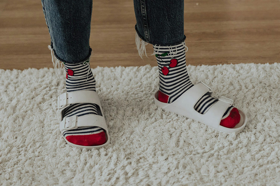 Two feet in black and white striped socks with red toes and heels in white slide sandals on white carpet.