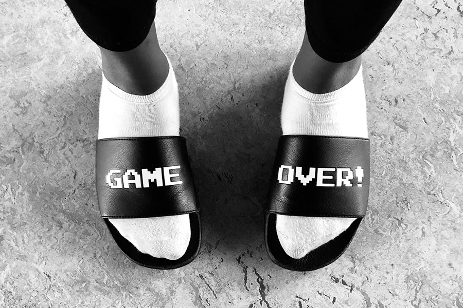 Two feet in white socks and black slide sandals with the word "Game" on one sandal and the word "Over!" on the other.