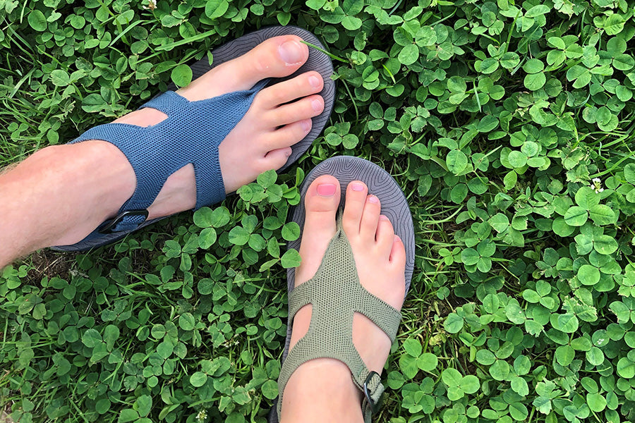 Two different female feet on a green background, one wearing a blue sandal, one wearing a green sandal.