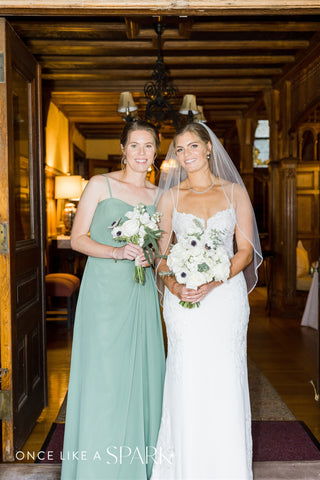 Image of bride and bridesmaid. Bride is holding her bridal bouquet with white and green florals. The bridesmaid is in a sage green dress holding a matching bouquet