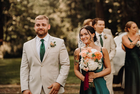 Image of bridesmaid and groomsmen walking down the aisle after the ceremony. The groomsman is in a light khaki color suit with green tie and pale pink boutonniere, the bridesmaid is in an emerald green long dress with a pale pink bridesmaid bouquet.